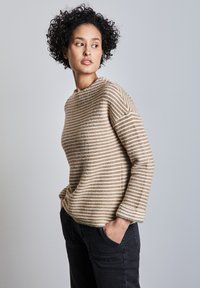 Beige gestreifter Pullover mit langen Ärmeln, weicher Textur, geripptem Halsauschnitt und lässiger Passform. Kombiniert mit schwarzen Hosen und Händen in den Taschen.