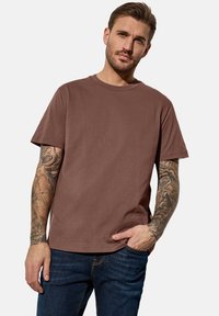 Hnědé bavlněné tričko s kruhovým výstřihem, krátkými rukávy a uvolněným střihem, v kombinaci s modrými džíny. Zobrazeny viditelné tetování na ruce.