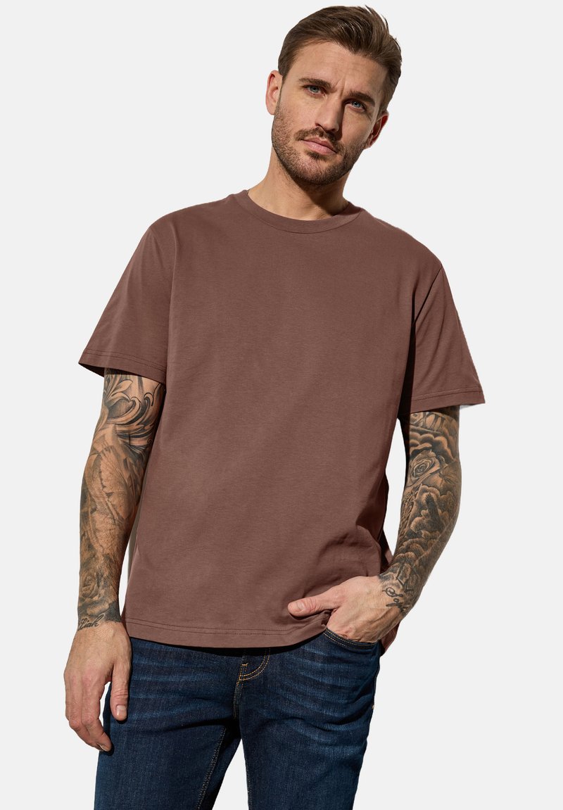 Hnědé bavlněné tričko s kruhovým výstřihem, krátkými rukávy a uvolněným střihem, v kombinaci s modrými džíny. Zobrazeny viditelné tetování na ruce.