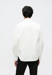 Camicia bianca a maniche lunghe in cotone, con colletto classico e polsini con bottoni, mostrata da dietro su uno sfondo neutro.