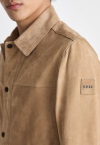 Chaqueta de ante color arena con cuello abotonado, que cuenta con un bolsillo en el pecho izquierdo y un parche con el logo "BOSS" en la manga.
