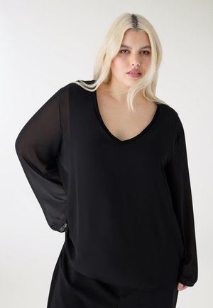 Donna bionda con capelli sciolti che indossa una camicetta nera a maniche lunghe con scollo a V e maniche trasparenti, in piedi davanti a uno sfondo bianco.