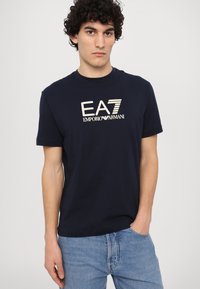 Temno modra bombažna majica z belim logotipom EA7 Emporio Armani čez prsi. Ima kratke rokave in krožni izrez.