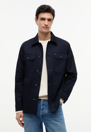 Mand iført en marineblå knapjakke med brystlommer over en cremefarvet sweater og blå jeans, stående mod en ensartet hvid baggrund.