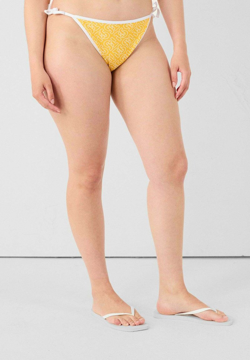 Gele bikinibroekjes met een geometrisch patroon, witte koordjes aan de zijkanten, gecombineerd met witte teenslippers op een neutrale achtergrond.