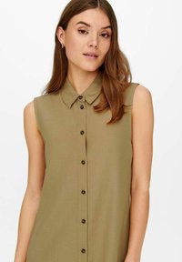 Licht olijfgroene mouwloze shirtjurk met een kraag, heeft een knoopsluiting aan de voorkant en een subtiele textuur. Geen patronen of accenten aanwezig.