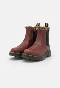 Botas de cuero marrón hasta el tobillo con cuello de pelaje sintético, paneles laterales elásticos, costuras amarillas y suelas de goma negra. Diseño robusto para uso casual.