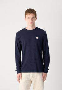 Marineblauwe longsleeve t-shirt van geweven stof met een ronde hals en een klein logo-embleem op de linkerborst. Voorzien van ribbels aan de mouwen.