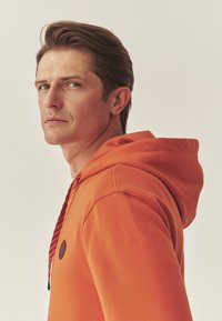 Oranje hoodie met een trekkoord en zwarte strepen. Het heeft een ronde, donkere patch op de borst en een zachte textuur.