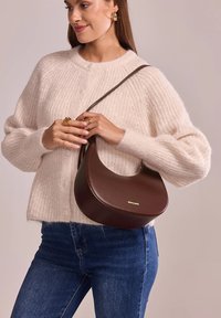 Sac à bandoulière en cuir marron avec une forme incurvée et un logo doré, porté avec un pull en tricot crème et un jean bleu.