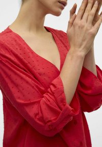 Blusa rossa con scollatura a V, tessuto testurizzato con punti in rilievo e maniche arrotolate con dettagli in bottone. Materiale morbido e leggero.