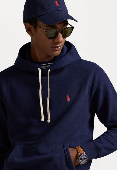 Sudadera navy con un bolsillo frontal y cordones crema. Presenta un logo rojo en el pecho y una camiseta a rayas debajo. Reloj marrón en la muñeca.