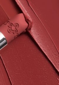 Rossetto opaco in una tonalità marrone-rossa calda, con una punta arrotondata e un involucro metallico argentato, applicato su una base liscia e testurizzata.