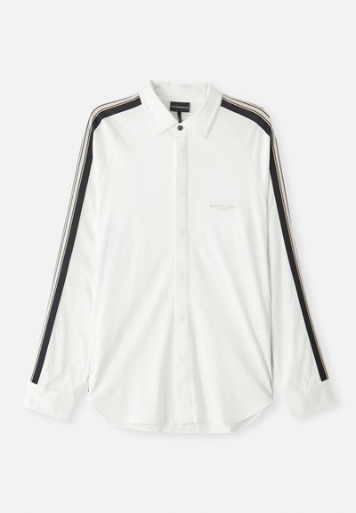 Balta pogu krekla ar melniem un bežiem svītrām uz piedurknēm, norādītas apkakles un izšūta zīmola logo uz krūtīm.