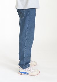 Jeans de mezclilla en azul medio con corte recto, que presentan un patrón de costura decorativa en el bolsillo trasero y una textura sutil.
