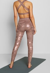 Bralette deportiva con tirantes entrecruzados en malva claro, combinada con leggings de cintura alta y largo a la espinilla que presentan un patrón de camuflaje brillante.