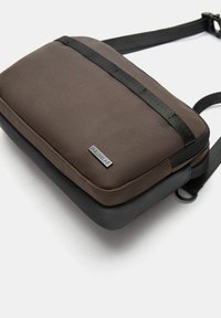 Braune, strukturierte Schultertasche mit einem schwarzen Riemen und einem glatten Boden, sowie einem kleinen metallischen Logo auf der Vorderseite.