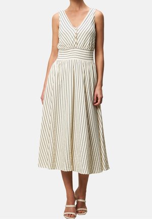 Ærmeløs beige mellemlang kjole med lodrette sorte striber, V-hals med knapper, tætsiddende taljebånd og plisseret nederdel, båret med beige hæle.
