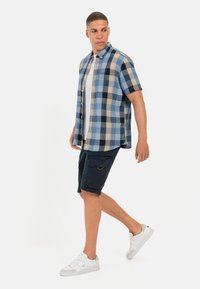 Kortekooien shirt in blauw en beige, met knopen, gecombineerd met marineblauwe cargoshorts met zakken, en witte sneakers.