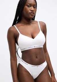 Biely bezšvový bralette s tenkými ramienkami a pásikom s logom; zodpovedajúce biele bikini nohavičky s pásikom s logom. Hladká textúra.