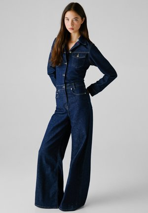 Combinaison en denim bleu foncé avec un haut ajusté, des pantalons à jambes larges, des boutons à l'avant et des poches sur la poitrine. Présente des manches longues et des coutures minimales.