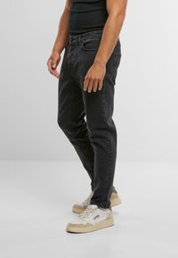 Jeans in denim nero con una vestibilità slim, cinque tasche e cuciture a contrasto; indossati con sneakers bianche con accenti beige e suola testurizzata.