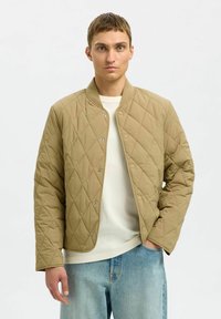 Pikowana beżowa kurtka bomberka z zatrzaskami, żebrowanym kołnierzem i długimi rękawami, noszona na jasnej koszuli i dżinsach.