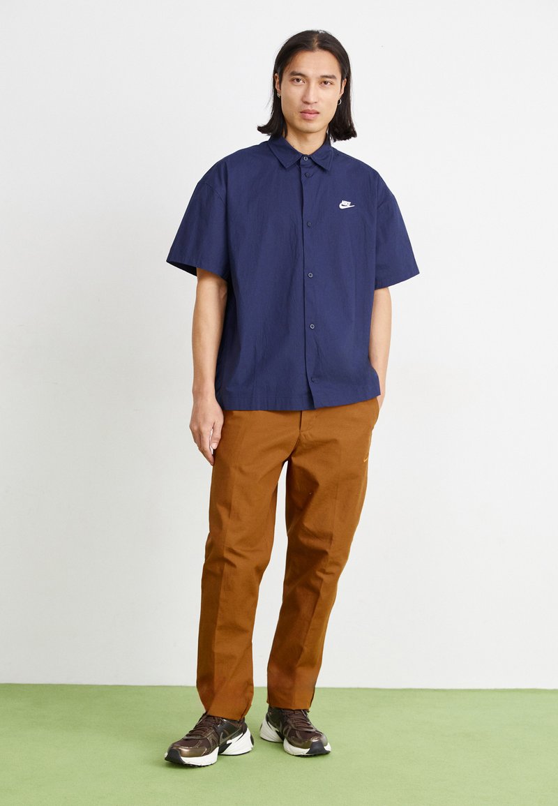 Chemise à manches courtes bleu marine avec un col et un logo blanc, assortie à un pantalon marron et des baskets foncées, sur un sol vert.