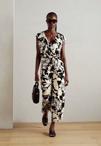 Jumpsuit floreale in bianco e nero con maniche corte, dettaglio con nodo frontale e gambe larghe, abbinato a sandali neri e una borsa.