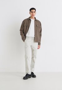 Brun skjortejakke over en hvit t-skjorte, sammen med lyse beige bukser og svarte snøresko, mot en nøytral bakgrunn.