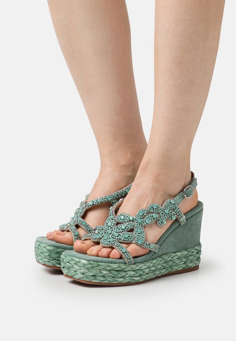 Pieds portant des sandales compensées vertes avec des semelles plateforme tressées et des brides ornées de bijoux en forme de fleurs, maintenues par des boucles à la cheville.
