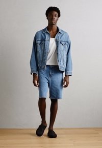 Denim jakna i kratke hlače svijetloplave boje, uparene s bijelom majicom na bubrege. Model nosi crne slip-on cipele. Prostorija ima svijetlo drveno pod.
