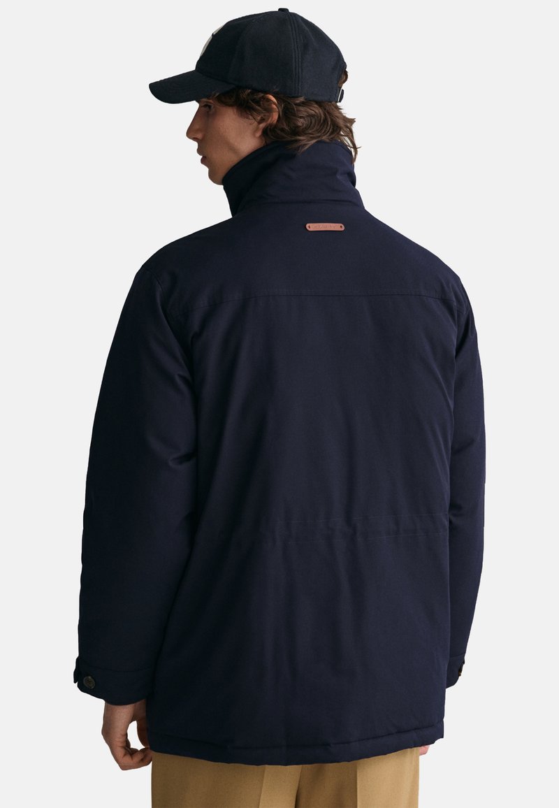 Giacca esterna blu navy con colletto alto, dettaglio in pelle sul retro e polsini con bottoni. Texture liscia, vestibilità rilassata e design di lunghezza media.