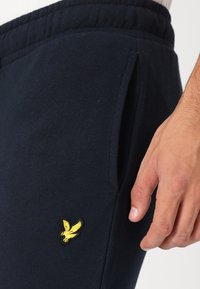 Temno modri trenirki z mehkimi teksturami, s stranskim žepom in rumenim vezenim logotipom orla na stegnu. Oblikovanje elastičnega pasu.