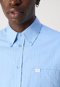 Lichtblauw geruit shirt met een button-down kraag, een borstzak en een klein label. Zachte textuur met een subtiel ruitpatroon.