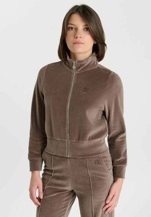 Femme aux cheveux courts et bruns porte une veste en velours marron zippée et un pantalon assorti, debout devant un fond blanc uni.
