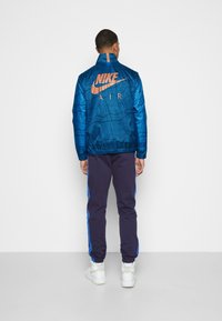 Sininen Nike-takki, jossa on abstrakti kuvio ja oranssi logo sekä säädettävä kaulus. Yhdistetty tummansinisiin housuihin, joissa on siniset sivuraidat.