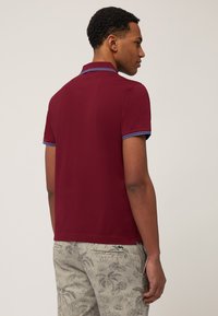 Burgundské polo tričko s krátkymi rukávmi, so modro pruhovaným lemovaním na golieri a rukávoch, skombinované s svetlými vzorovanými šortkami.