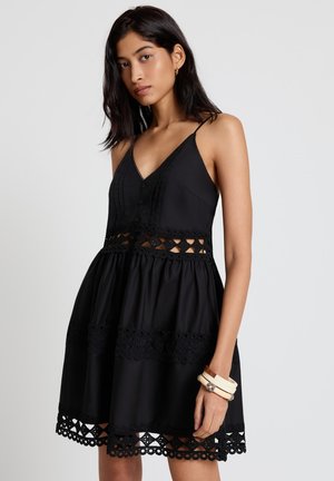 Giovane donna che indossa un abito nero senza maniche con intagli in pizzo e fasce decorative, arricchito da braccialetti bianchi al polso.