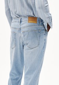 Jean en denim bleu clair avec une coupe droite, présentant une étiquette en cuir marron portant le nom "ARMEDANGELS" et des poches arrière visibles avec des détails de coutures.