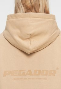 Béžová mikina s okrúhlou kapucňou, mäkkou textúrou a embosovaným logom "PEGADOR" na chrbte. Minimalistický dizajn s čistými líniami.