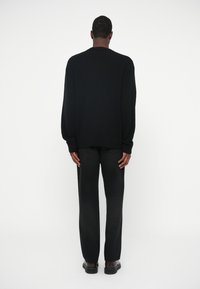 Pulover negru din tricot cu manșete cu nervuri, purtat cu blugi întunecați. Modelul stă cu spatele, pe un fundal alb simplu.