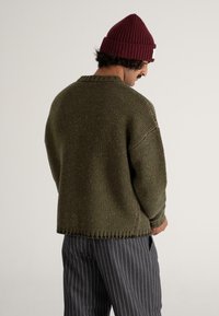 Green knitted sweater with a textured finish and a ribbed collar, paired with gray pinstripe pants, viewed from the back.