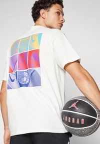 White cotton t-shirt featuring a multicolored grid design on the back with various basketball-related graphics; model holds a black basketball.