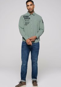 Lichtgroene button-up shirt met zwarte tekstontwerpen, één pocket, gecombineerd met blauwe spijkerbroek en grijsbruine schoenen.