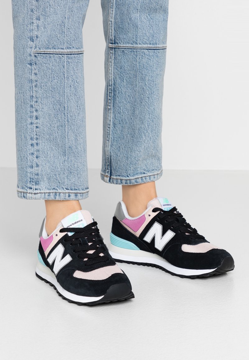 Baskets noires en daim et en maille avec des accents roses et bleu clair, arborant un logo "N" blanc et des lacets noirs, posées sur un jean en denim clair.