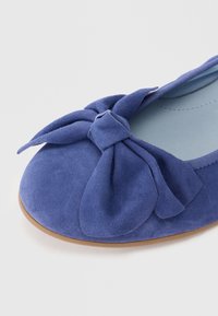 Fleur de ballet en daim bleu avec un nœud proéminent à l'avant, une doublure lisse et une semelle marron clair. Vue en gros plan.