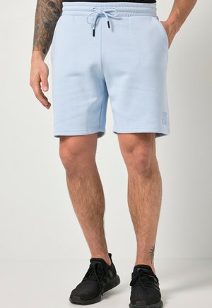 Mand iført lyseblå snoretrusser og sorte sneakers står med den ene hånd i lommen mod en ensfarvet baggrund.
