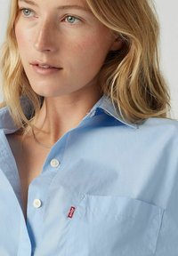 Camicia button-up azzurro chiaro con colletto, dotata di taschino sul petto e una piccola etichetta rossa. Il tessuto è liscio e leggermente lucido.