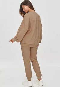 Brązowa, oversize'owa bluza i dopasowane dresy wykonane z miękkiego materiału. Posiadają ribbowe mankiety oraz białe sneakersy. Model pokazany z tyłu.